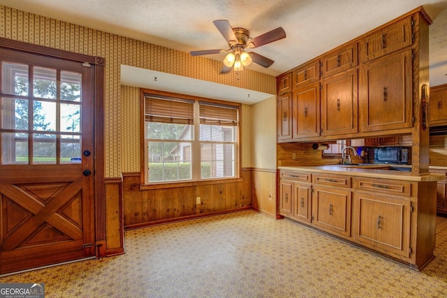 kitchen featuring wallpapered walls, black microwave, brown cabinetry, and wainscoting