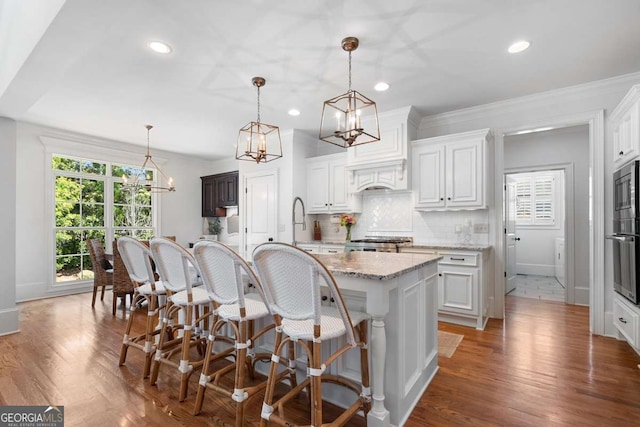 kitchen with range, white cabinetry, a kitchen island with sink, light stone counters, and decorative light fixtures