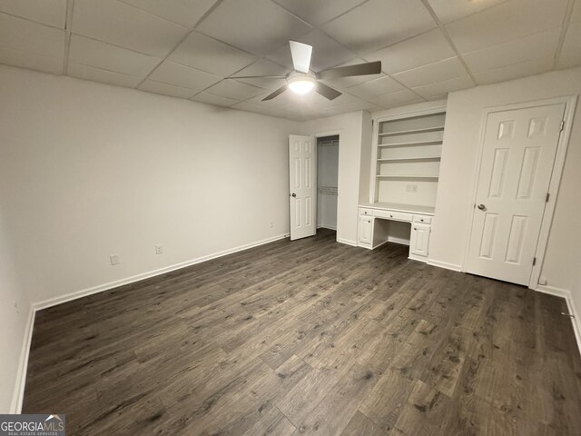 unfurnished bedroom with a drop ceiling, built in desk, dark hardwood / wood-style floors, and ceiling fan