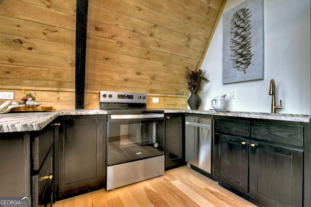 kitchen with stainless steel appliances, light wood-style floors, wooden walls, light stone countertops, and dark cabinets