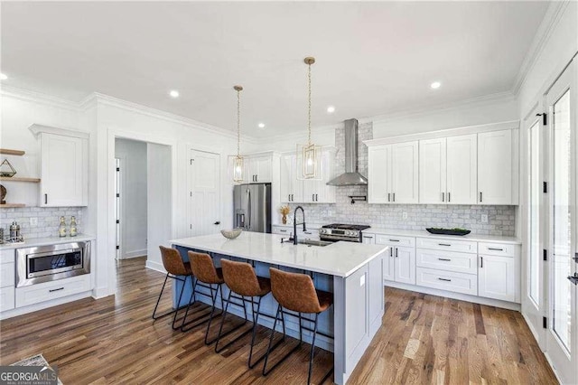 kitchen featuring white cabinetry, sink, wall chimney range hood, stainless steel appliances, and a center island with sink