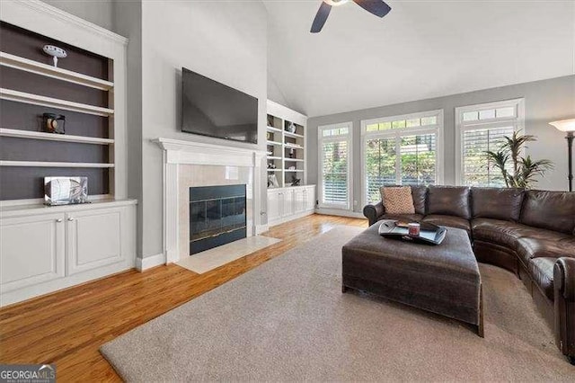 living room with built in features, lofted ceiling, a tiled fireplace, ceiling fan, and light hardwood / wood-style floors