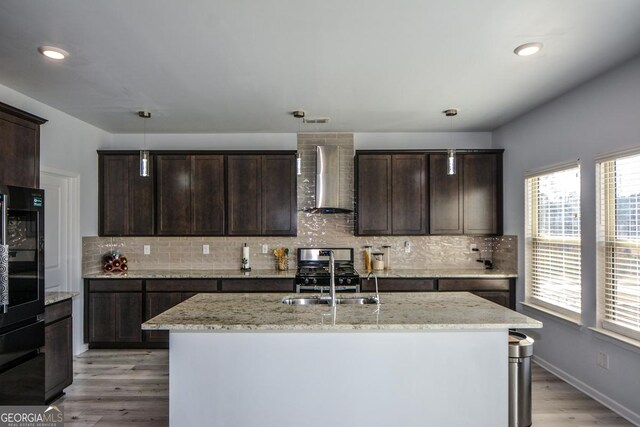 kitchen with sink, light stone counters, decorative light fixtures, dark brown cabinets, and wall chimney range hood