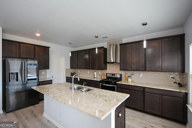 kitchen featuring sink, appliances with stainless steel finishes, an island with sink, pendant lighting, and wall chimney range hood