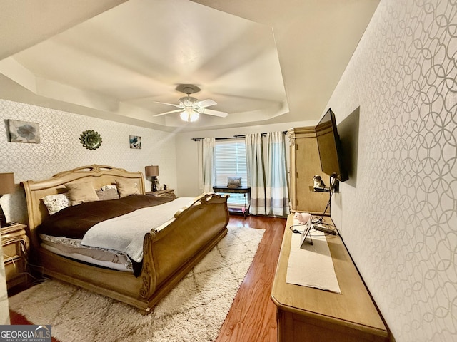 bedroom featuring ceiling fan, a raised ceiling, wood finished floors, and wallpapered walls