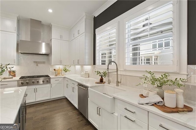 kitchen featuring wall chimney exhaust hood, sink, white cabinetry, light stone counters, and appliances with stainless steel finishes