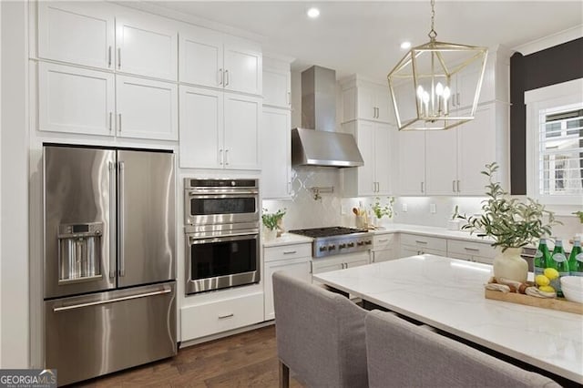 kitchen featuring stainless steel appliances, light stone countertops, white cabinets, decorative light fixtures, and wall chimney exhaust hood