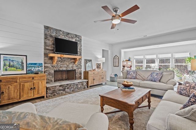 living room featuring wooden walls, ceiling fan, a stone fireplace, and light wood-type flooring