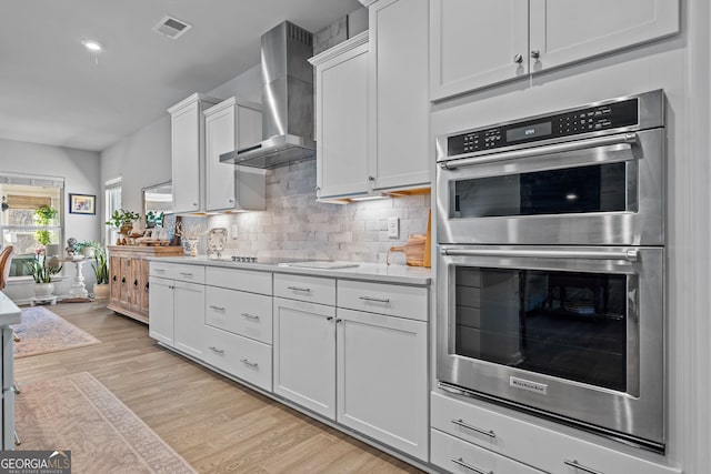 kitchen featuring light wood-type flooring, double oven, wall chimney range hood, decorative backsplash, and white cabinets