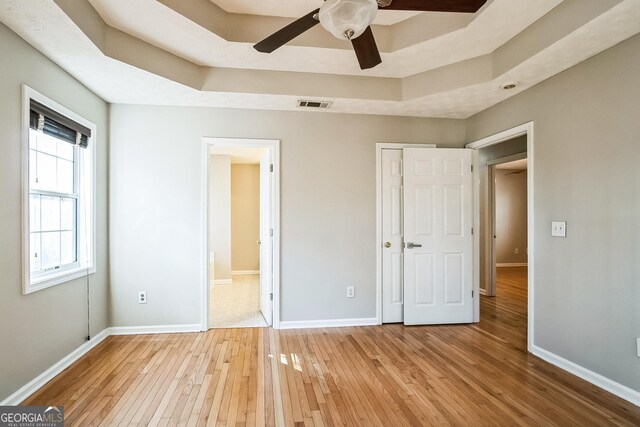 unfurnished bedroom with ceiling fan, ensuite bath, a raised ceiling, and light wood-type flooring