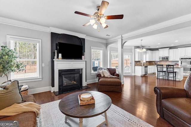 living area featuring ornamental molding, a glass covered fireplace, dark wood-type flooring, a ceiling fan, and baseboards