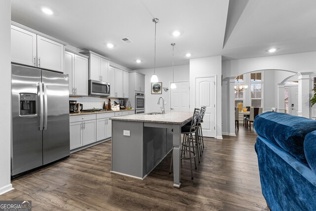 kitchen with appliances with stainless steel finishes, a kitchen island with sink, light stone countertops, white cabinets, and decorative light fixtures