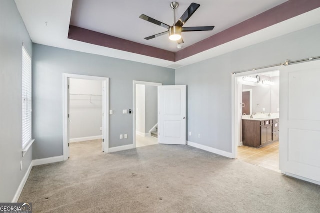 unfurnished bedroom featuring ensuite bathroom, a walk in closet, light colored carpet, a raised ceiling, and a barn door