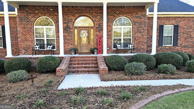 doorway to property featuring a porch, a shingled roof, and brick siding