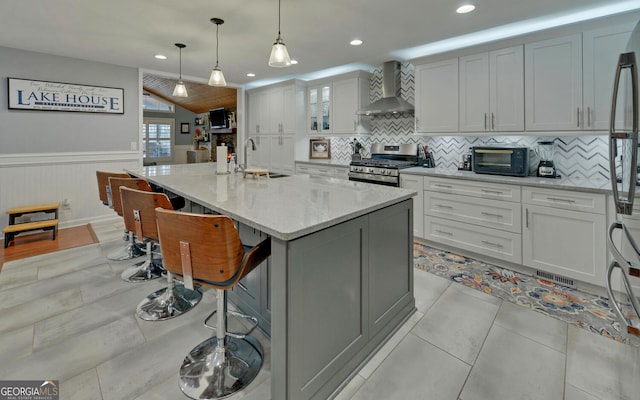 kitchen featuring wall chimney range hood, sink, a kitchen island with sink, light stone counters, and stainless steel range with gas cooktop