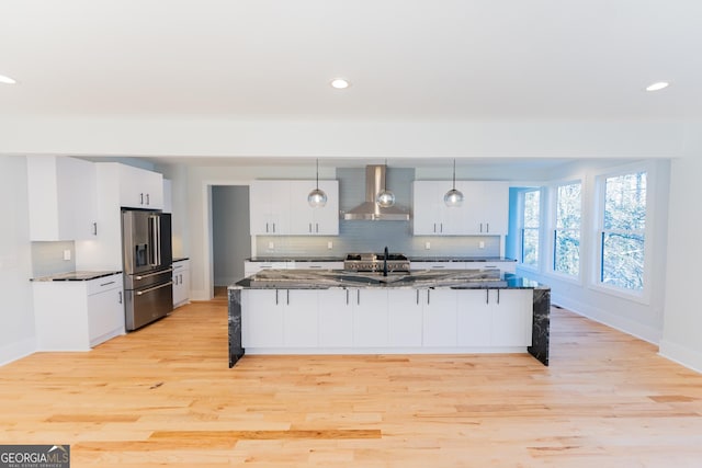 kitchen with wall chimney range hood, high end fridge, hanging light fixtures, dark stone countertops, and white cabinets