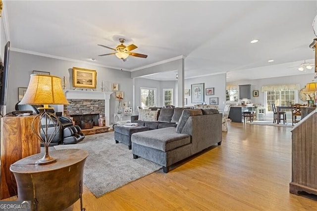 living room featuring ceiling fan, ornamental molding, a stone fireplace, and light wood-type flooring