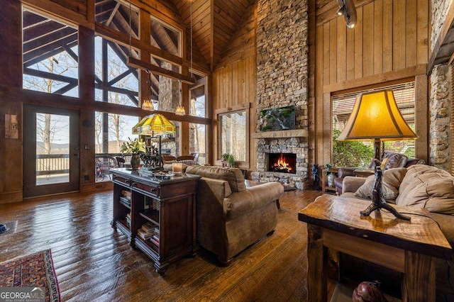 living room featuring a stone fireplace, rail lighting, high vaulted ceiling, wooden walls, and dark hardwood / wood-style floors