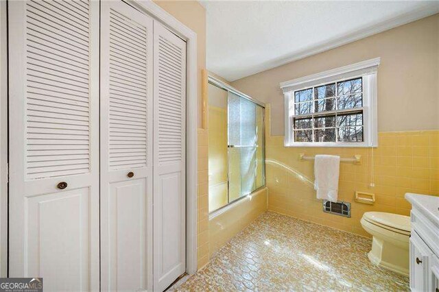 full bathroom featuring bath / shower combo with glass door, vanity, toilet, and tile walls