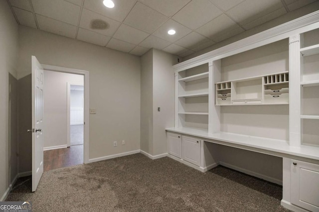 unfurnished office featuring a paneled ceiling, built in desk, and dark carpet