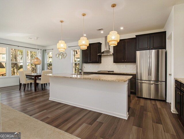 kitchen featuring pendant lighting, stainless steel appliances, a center island with sink, and wall chimney range hood