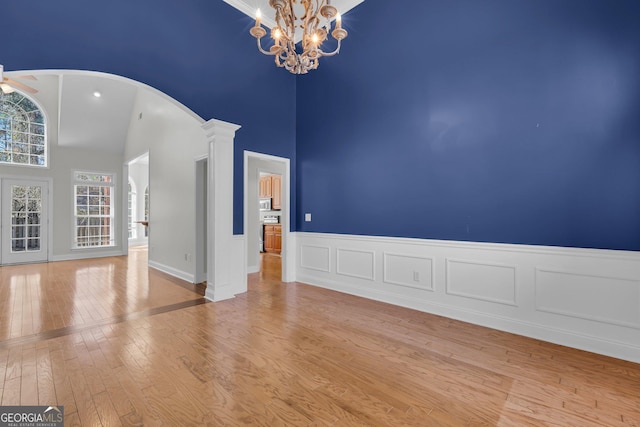 unfurnished room featuring a high ceiling, ceiling fan with notable chandelier, decorative columns, and light wood-type flooring