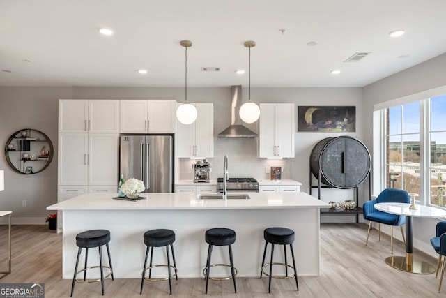 kitchen with white cabinetry, a center island with sink, high quality fridge, pendant lighting, and wall chimney range hood