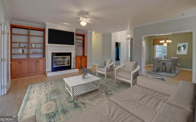 living area with ceiling fan with notable chandelier, baseboards, light wood finished floors, a glass covered fireplace, and crown molding