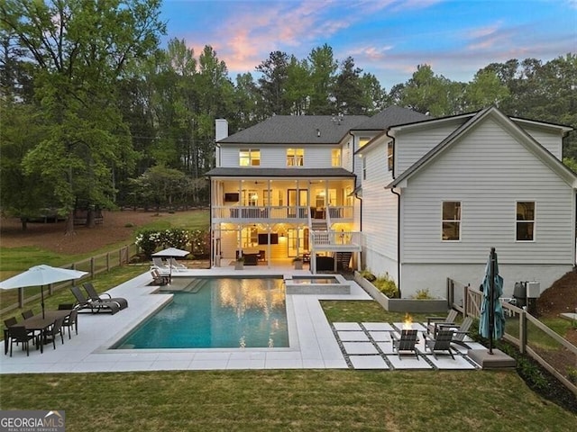 back house at dusk with a yard, a patio, an outdoor fire pit, a pool with hot tub, and a balcony