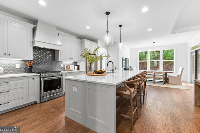 kitchen featuring a breakfast bar, stainless steel gas stove, custom range hood, an island with sink, and decorative light fixtures