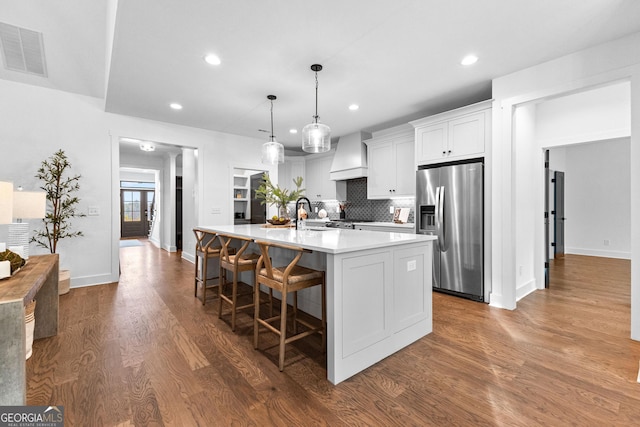 kitchen with premium range hood, pendant lighting, white cabinets, stainless steel refrigerator with ice dispenser, and a center island with sink