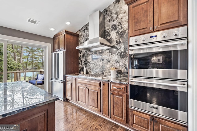 kitchen featuring wall chimney range hood, dark stone countertops, stainless steel appliances, dark hardwood / wood-style flooring, and decorative backsplash