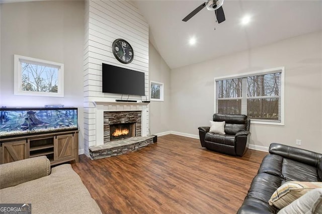 living area featuring baseboards, ceiling fan, wood finished floors, a stone fireplace, and high vaulted ceiling