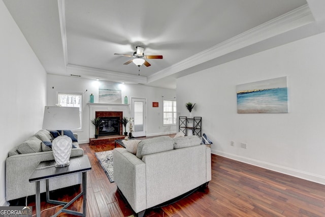 living room with dark hardwood / wood-style flooring, ornamental molding, ceiling fan, a tray ceiling, and a brick fireplace