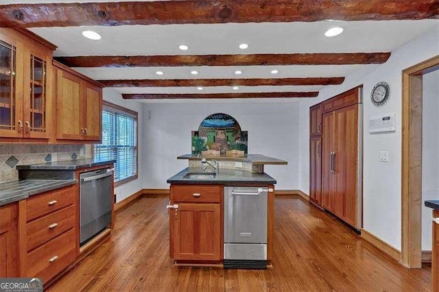 kitchen featuring wood-type flooring, a center island with sink, dishwasher, beamed ceiling, and decorative backsplash