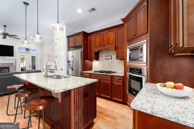 kitchen with sink, a kitchen island with sink, stainless steel appliances, light stone countertops, and decorative light fixtures