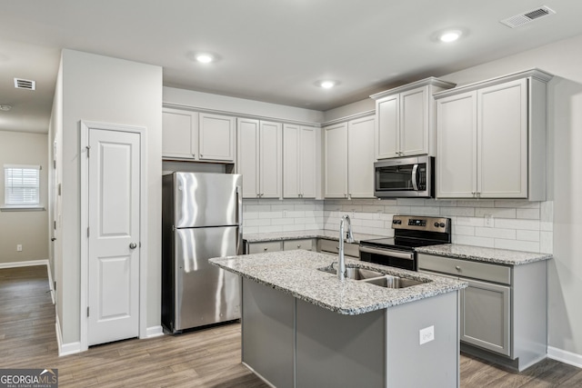 kitchen featuring appliances with stainless steel finishes, light stone countertops, light hardwood / wood-style floors, a center island with sink, and decorative backsplash