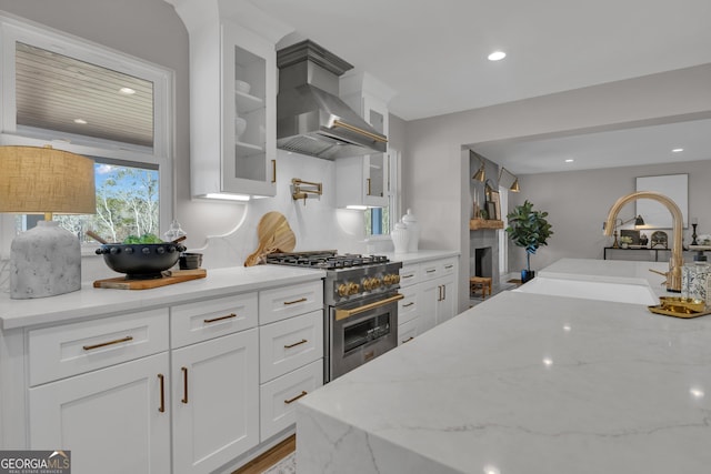 kitchen with sink, high end range, white cabinetry, ventilation hood, and light stone countertops