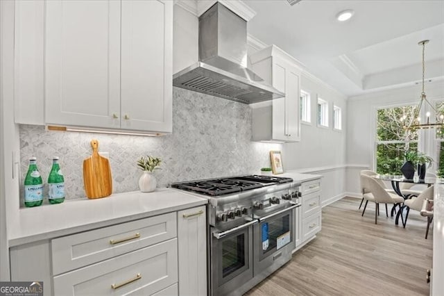 kitchen featuring pendant lighting, white cabinets, range with two ovens, a raised ceiling, and wall chimney range hood