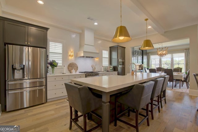 kitchen with custom exhaust hood, hanging light fixtures, appliances with stainless steel finishes, a kitchen breakfast bar, and a kitchen island with sink