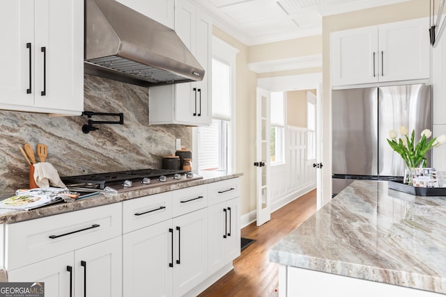 kitchen with ventilation hood, ornamental molding, stainless steel appliances, wood finished floors, and white cabinetry