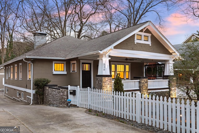 view of front of home featuring a ceiling fan, a porch, a shingled roof, a chimney, and a fenced front yard