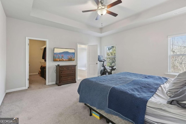 bedroom with a walk in closet, light colored carpet, ceiling fan, a raised ceiling, and ensuite bath