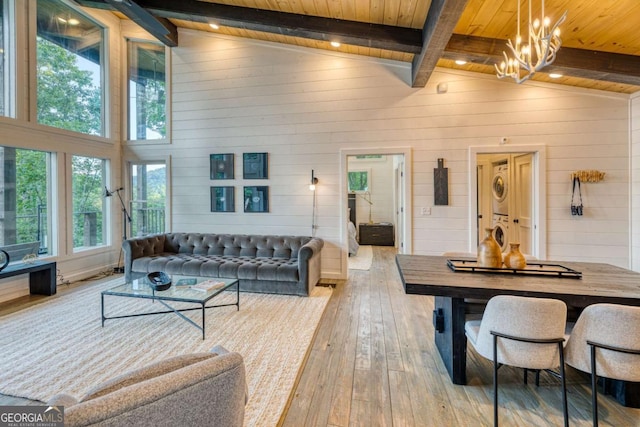 living room featuring light hardwood / wood-style flooring, stacked washing maching and dryer, a notable chandelier, wooden ceiling, and beamed ceiling