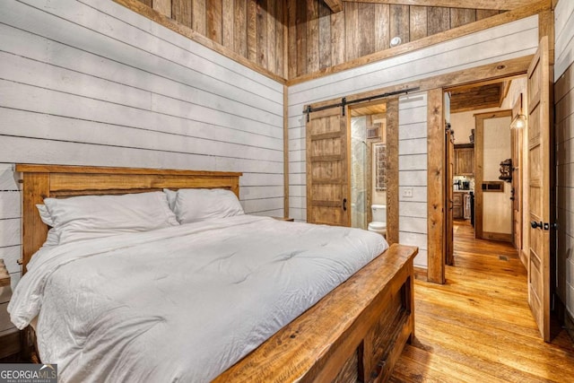 bedroom with ensuite bathroom, a barn door, wooden walls, and light hardwood / wood-style flooring