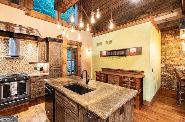 kitchen with wall chimney exhaust hood, sink, stainless steel gas range oven, hanging light fixtures, and an island with sink