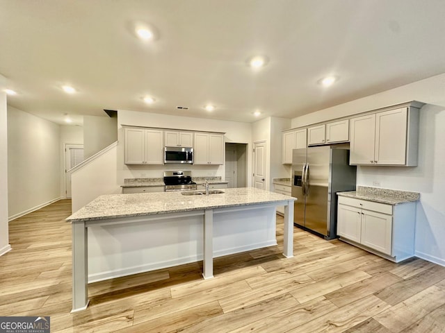 kitchen featuring sink, stainless steel appliances, light stone counters, a center island with sink, and light wood-type flooring