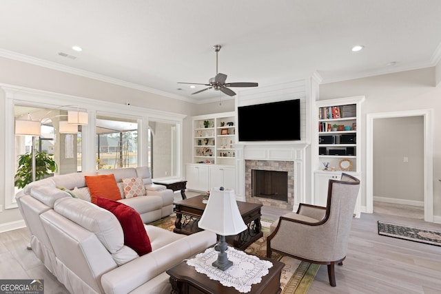 living room with a stone fireplace, built in features, ceiling fan, light hardwood / wood-style floors, and crown molding