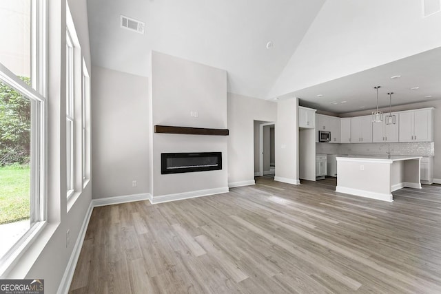 unfurnished living room featuring high vaulted ceiling, a sink, visible vents, light wood-type flooring, and a glass covered fireplace