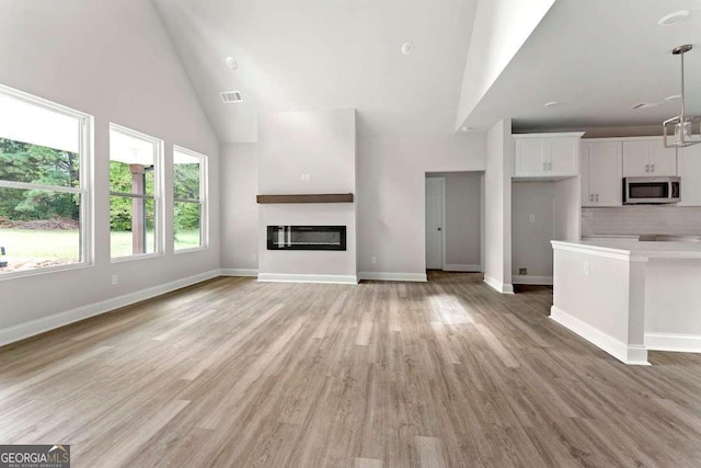 unfurnished living room featuring baseboards, visible vents, a glass covered fireplace, light wood-type flooring, and high vaulted ceiling